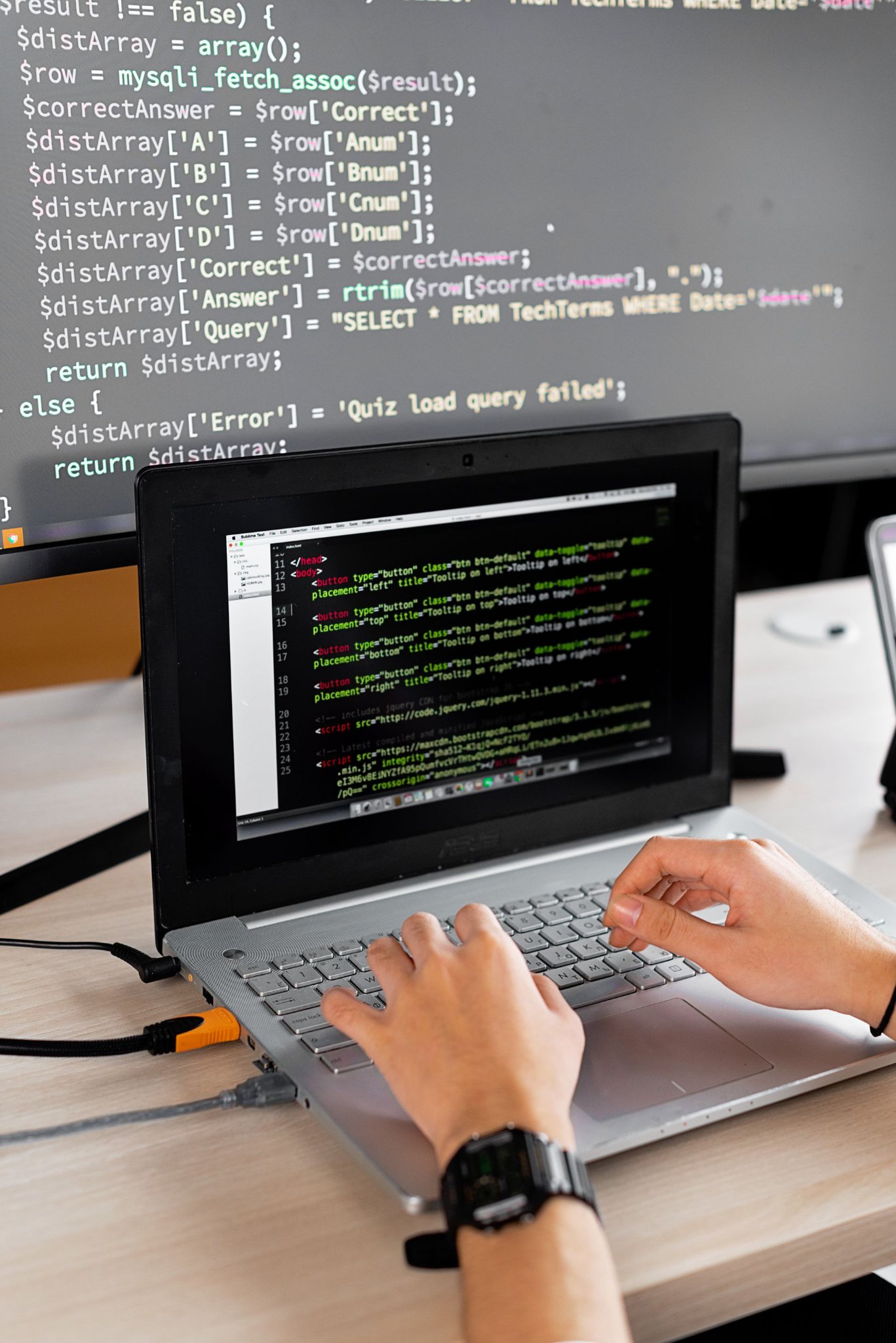 Photo of a person typing code on a laptop, with code on a large screen behind it
